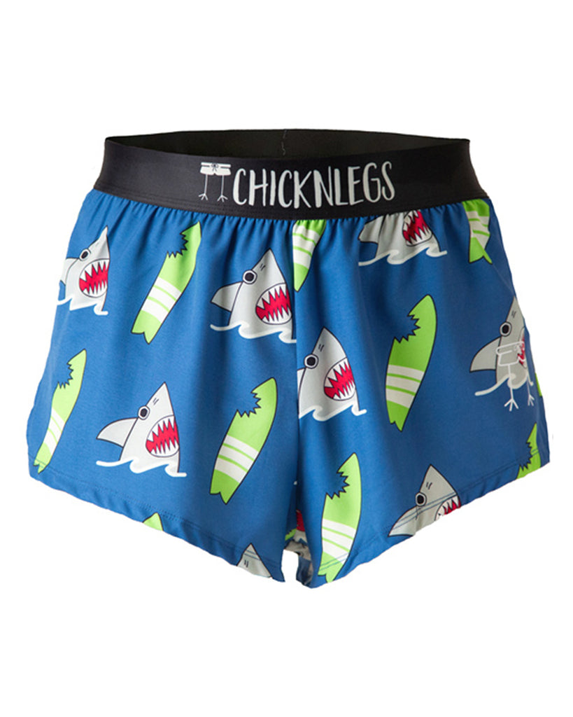 Chicknlegs men's 2 in split running shorts in the blue sharks design, as a ghost image. The print has a blue background with green surfboards with bite marks on it with a grey gray shark coming out of the water. These shorts are made for runners.