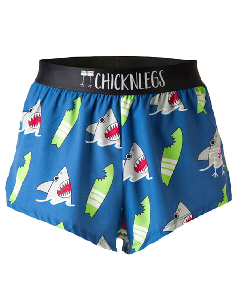 Chicknlegs men's 2 in split running shorts in the blue sharks design, as a ghost image. The print has a blue background with green surfboards with bite marks on it with a grey gray shark coming out of the water. These shorts are made for runners.