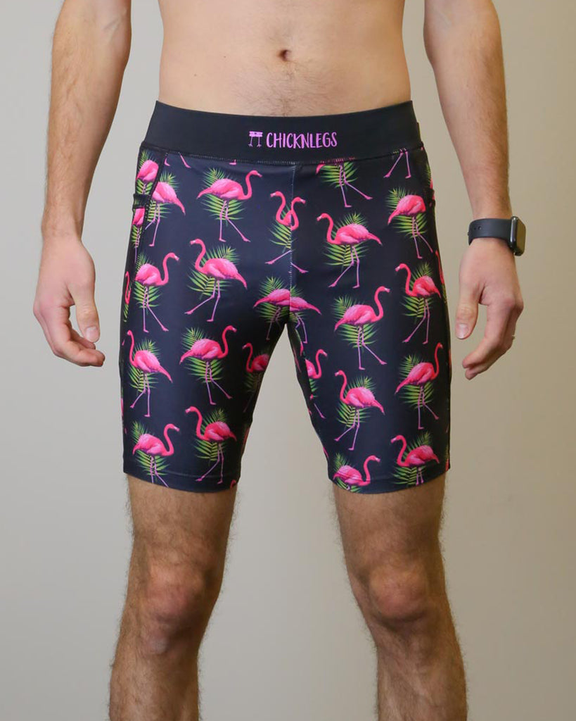 Runner is wearing ChicknLegs in the men's 8 in half tights with pockets in the flamingo design. The print has fun pink flamingos on it with green leaves on a solid black background.