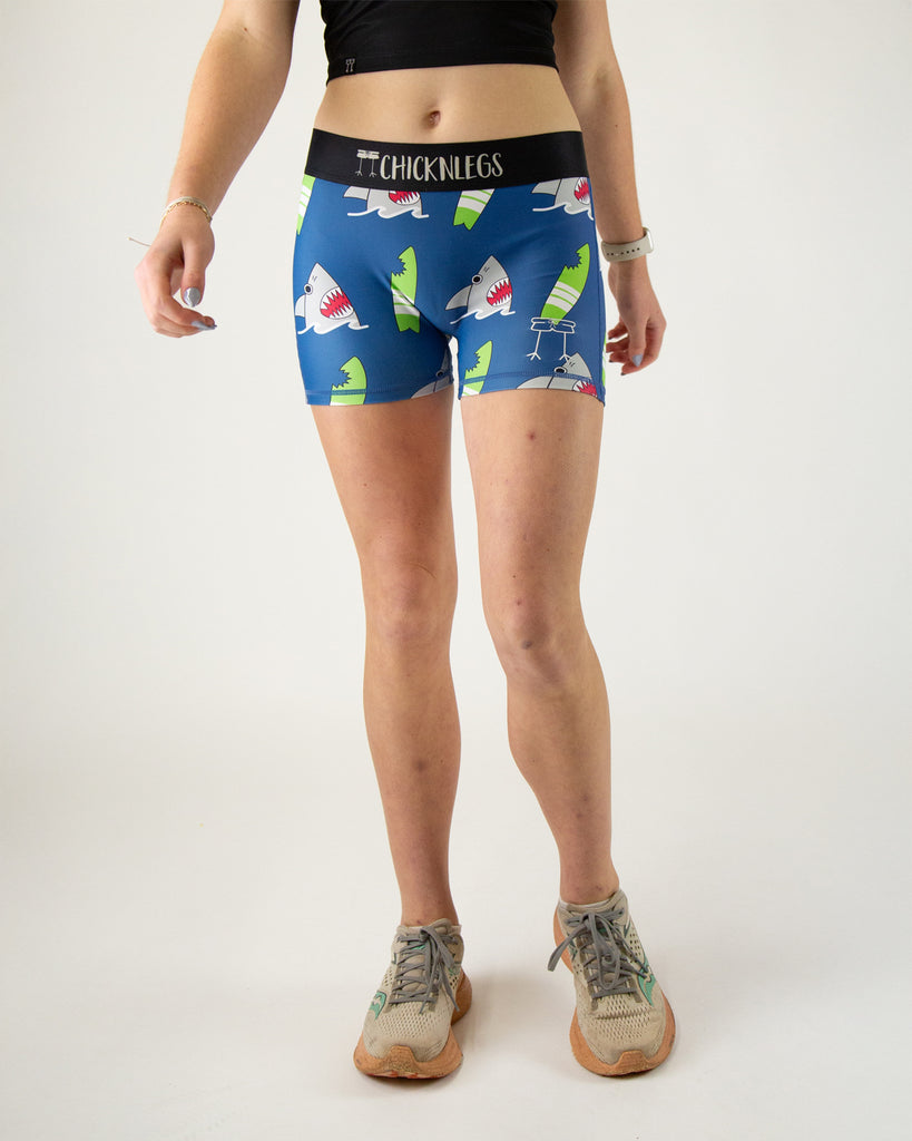 Runner is wearing ChicknLegs in the women's 3 in compression spandex shorts with the blue sharks design. The print has grey gray sharks on it with a green surfboard and a blue background.