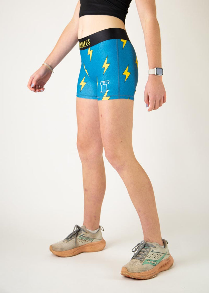 The model is a runner who is wearing Chicknlegs in the women's 3" compression shorts in the blue bolts design, facing left.