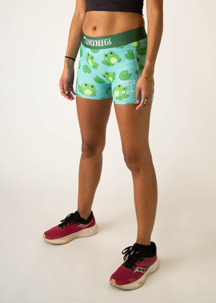 Model is a runner wearing Chicknlegs 3 inch compression shorts in the fartlek froggies frog design, facing left.
