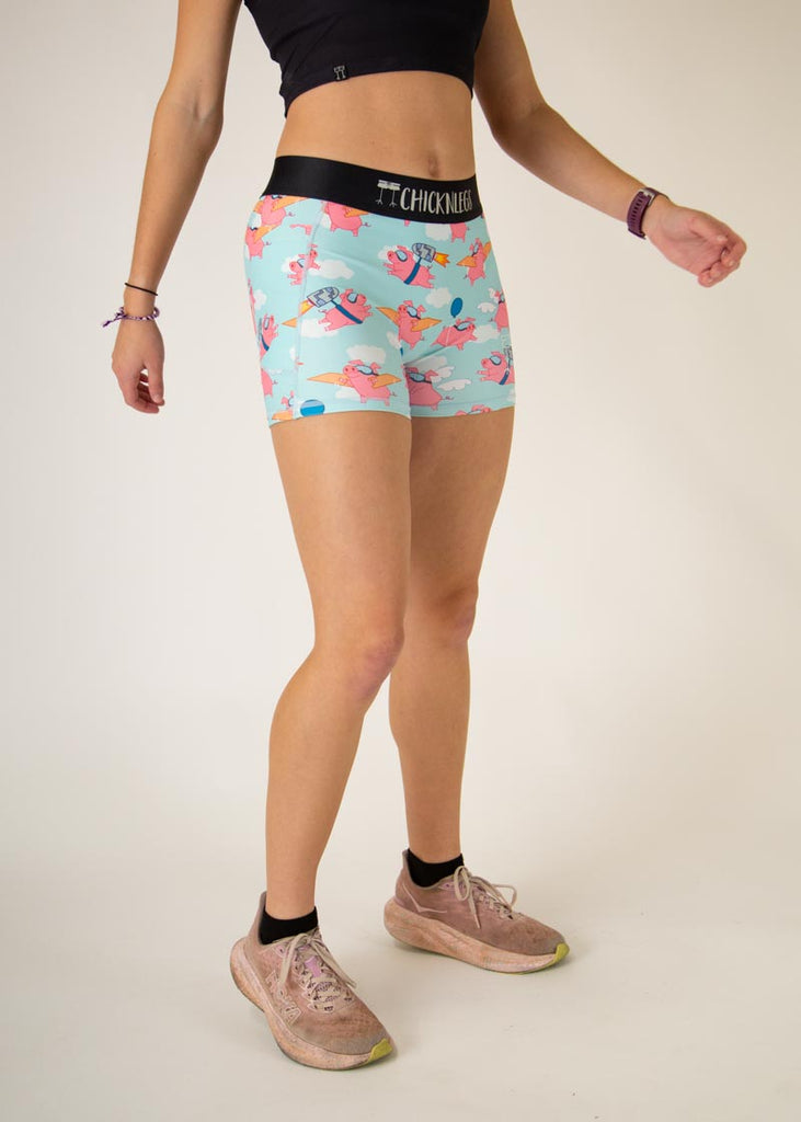 Runner is wearing ChicknLegs in the women's 3 in compression spandex shorts with side panel pockets that fit high rise on the waist. The design has pink piggies on it with cardboard wings, jetpacks and goggles with white clouds on a blue background.