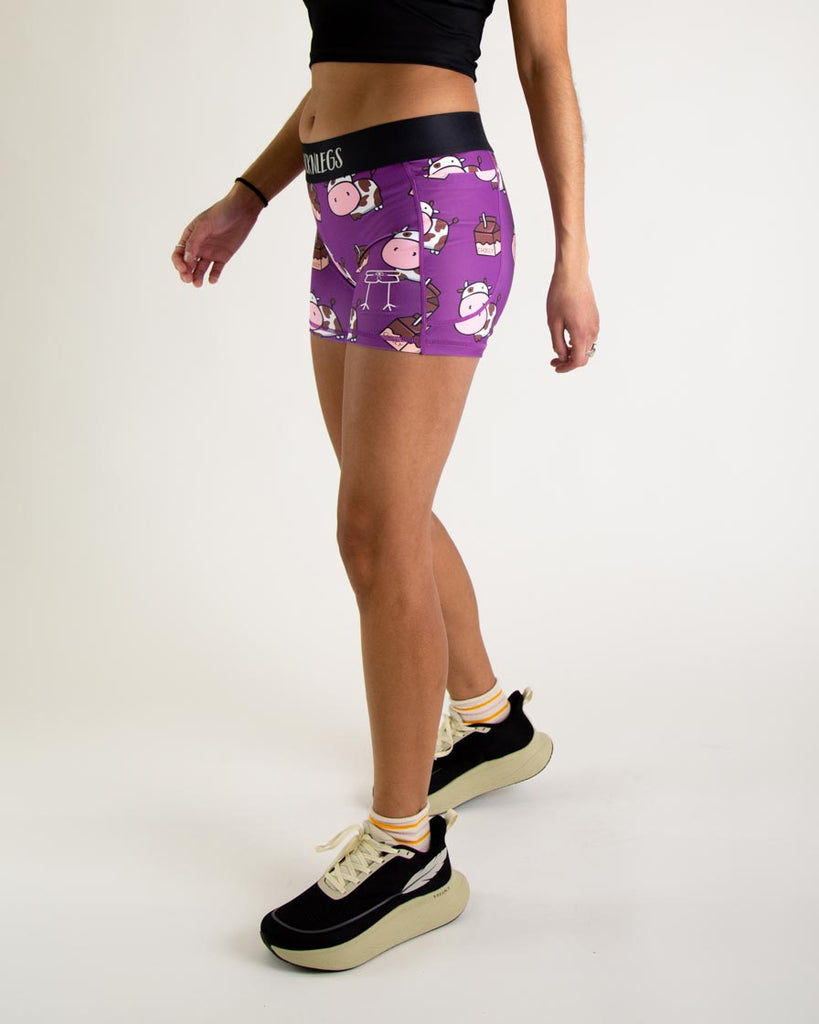 Runner is wearing ChicknLegs in the women's 3 in compression spandex shorts with pocket in the choccy cows design. The print has brown and white chocolate cows with milk cartons on it. The model is facing left.