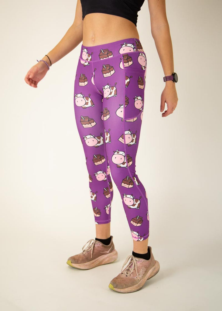 The model is a runner who is wearing Chicknlegs Women's Leggings in the Choccy Cow design. These leggings include side pockets and an adjustable drawstring at the front. The model is facing slightly left.