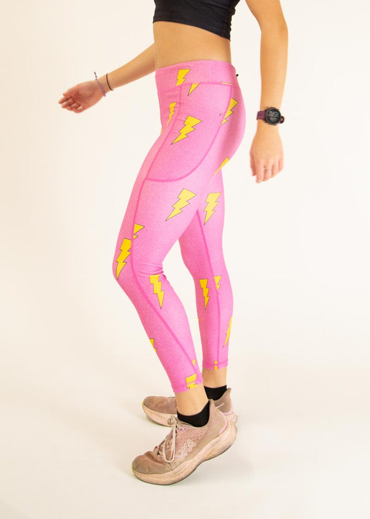 The model is a runner who is wearing Chicknlegs Women's Leggings in the Hot Pink Bolts design. These leggings include side pockets and an adjustable drawstring at the front. The model is facing left to show the side view.