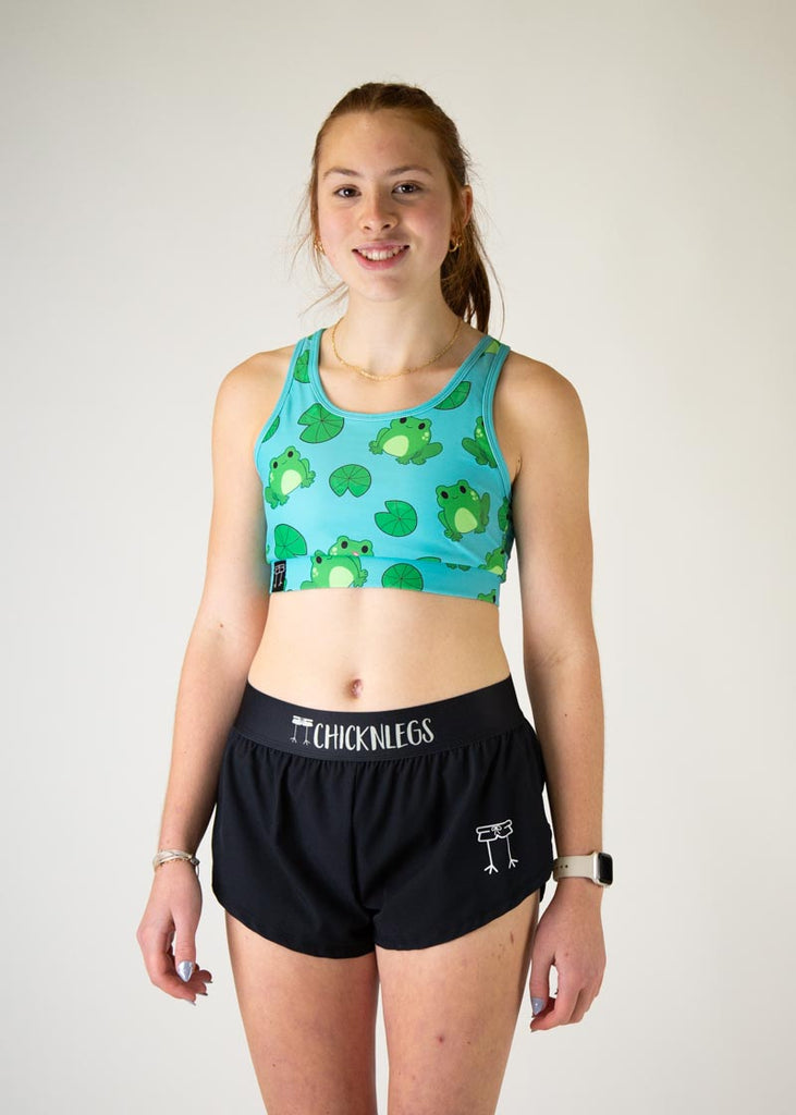 The model is a runner who is wearing Chicknlegs in the women's OG sports bra, in the fartlek froggies frog design, facing front. 