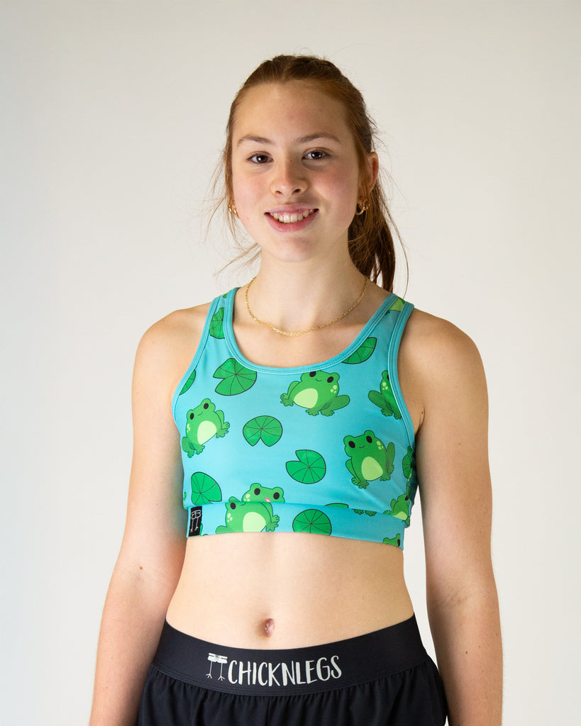 Runner is wearing ChicknLegs in the women's OG Sports Bra has a back pocket for exercise, running, workout, gym, and every day lifestyle. The print is the fartlek froggies design which has green frogs frog and lilypads on a solid blue background.