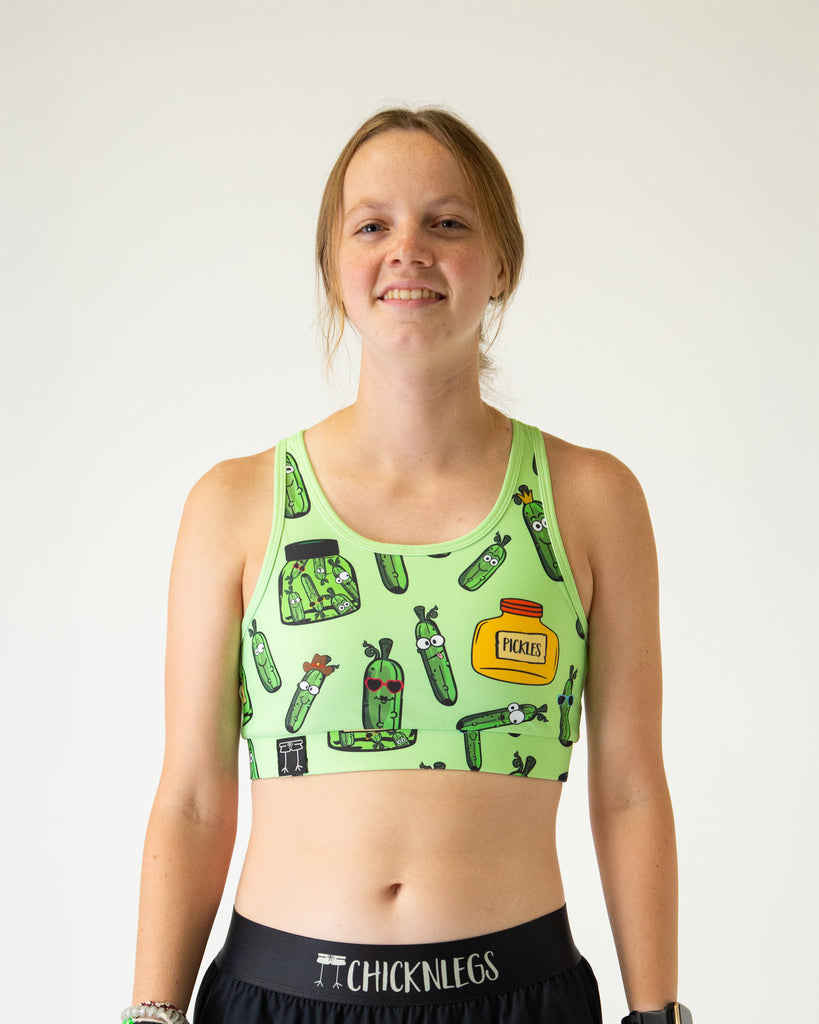 Runner is wearing ChicknLegs in the women's OG Sports Bra has a back pocket for exercise, running, workout, gym, and every day lifestyle. The print is the pickles design which has green pickles in different shapes and sizes and a green and yellow jars.