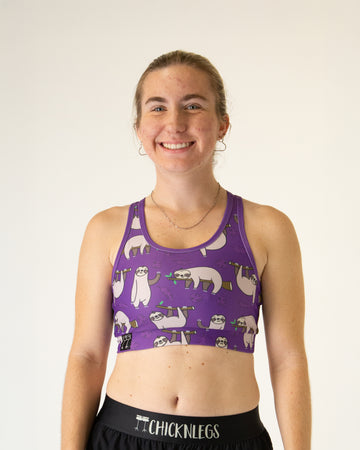 Runner is wearing ChicknLegs in the women's OG Sports Bra has a back pocket for exercise, running, workout, gym, and every day lifestyle. The print is the sloth design which has many sloths standing and sleeping with tree branches and leaves on a purple background.