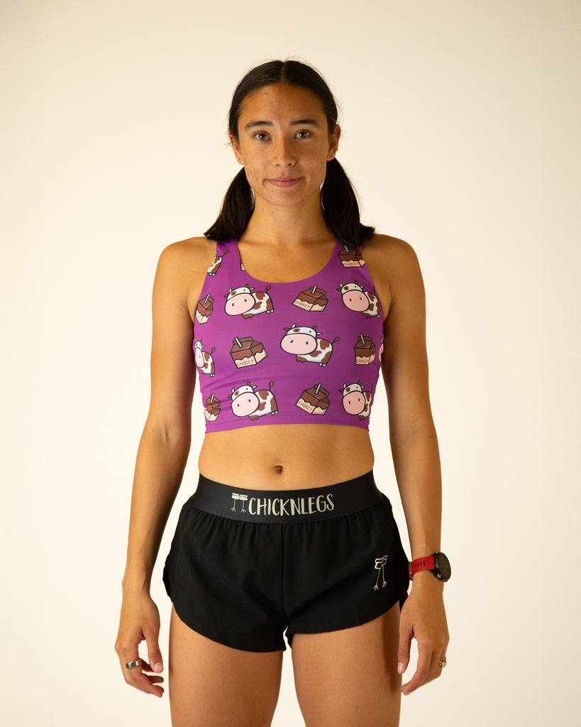 Runner is wearing ChicknLegs in the women's power crop top in the choccy cow design. The material is lightweight and the print has brown and white cows on it with chocolate milk on a purple background.