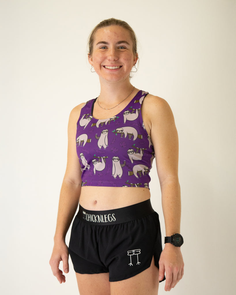 Runner is wearing ChicknLegs in the women's power crop top in the sloths design. The material is lightweight and the print has sloths with branches and leaves with a solid purple background.